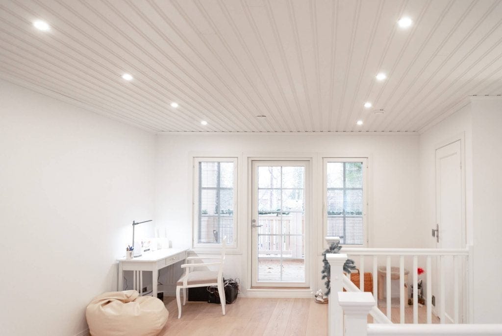 Valoisa ja minimalistinen huone, jossa på valkoiset seinät ja katto. Vasemmalla puolella sijaitsee pöytä ja tuoli, ja tilassa on myös säkkituoli sekä ovi, joka johtaa pienelle parvekkeelle. Upotetut LED-valot valaisevat tillan kauniisti. Kulmassa seisoo pieni joulukuusi.