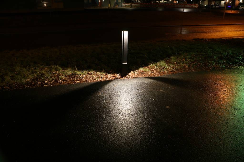 Hämärällä kadulla yössä seisoo LedStore.fi:n laadukas LED-valaisin, joka heijastaa kirkkaan kolmiomaisen valon märälle jalkakäytävälle. Ympäröivä alue pysyy pimeänä, mutta valaistus tuo esiin heikkoja vihjeitä ruohosta ja pudonneista lehdistä.