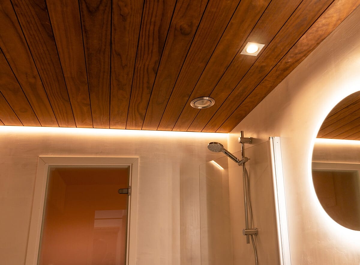 Modernissa kylpyhuoneessa på puupaneloitu katto, johon på upotettu LED-valaistus LedStore.fi:stä. Seinää koristaa pyöreä peili taustavalolla, joka sijaitsee tyylikkään suihkupään vieressä. Tämä luo tilaan lämpimän ja kutsuvan tunnelman.