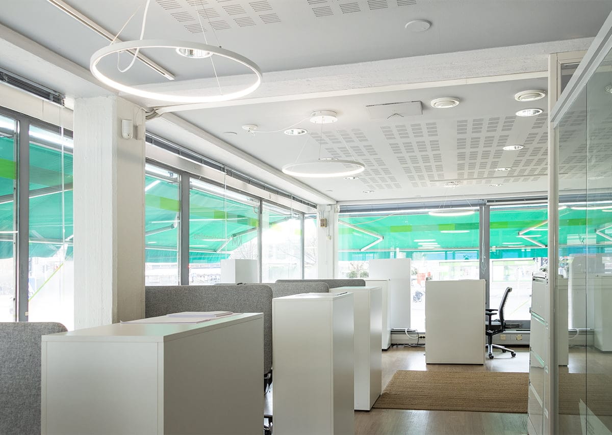 LedStore.fi tarjoaa moderniin ja valoisan toimistotilan, jossa på valkoiset työpöydät ja harmaat väliseinät. Suuret ikkunat vihrein markiisein päästävät runsaasti luonnonvaloa sisään. Huoneen valaisee tehokkaasti LED-kattovalaisimet, kun taas osa puulattiasta på peitetty beigenvärisellä matolla. Näkyvissä på myös toimistotuoli.