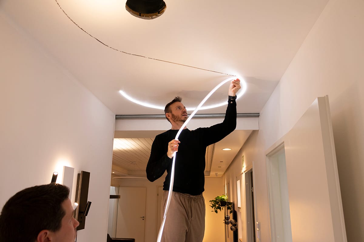 Person på stege installerar ljus LED-remsa längs taket; rum väl upplyst. Observer nedan ser som flexibel LED döljer synliga taksprickor.