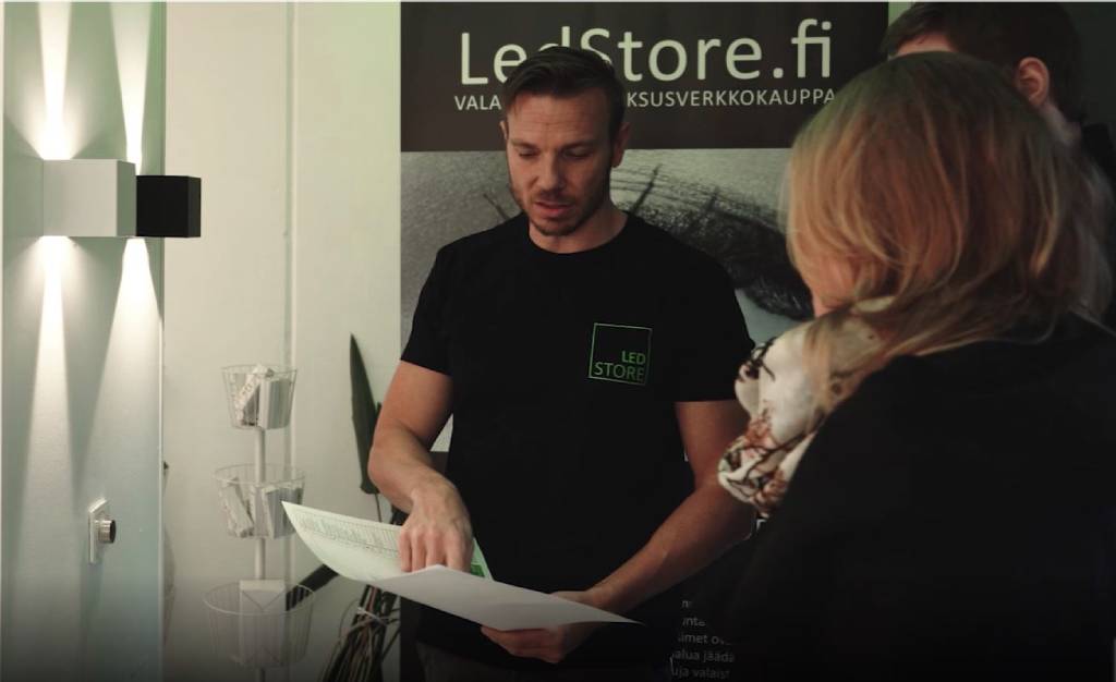 LedStore.fi-verkkokaupan pehmeästi valaistussa tilassa henkilö mustassa paidassa, jossa på LedStore-logo, pitää kädessään paperia ja selittää kahdelle henkilölle LED-valaistusteknologiaa. Taustalla näkyy "LedStore.fi" -banneri.