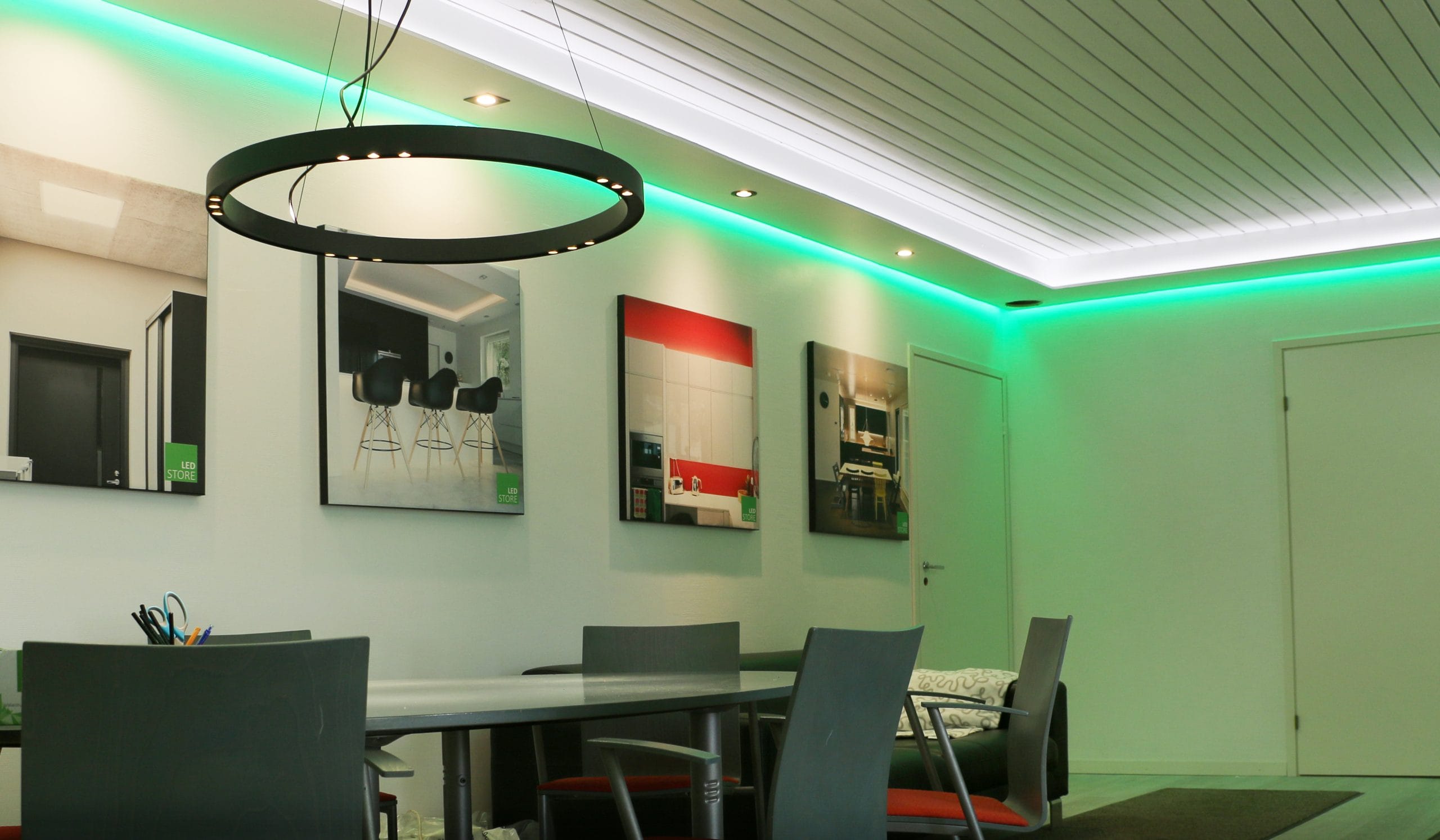 Modernt kontor med rund LED-armatur, grå stolar runt bordet, infällda taklampor, gröna LED-accenter, konstväggar, dörr och hylla.