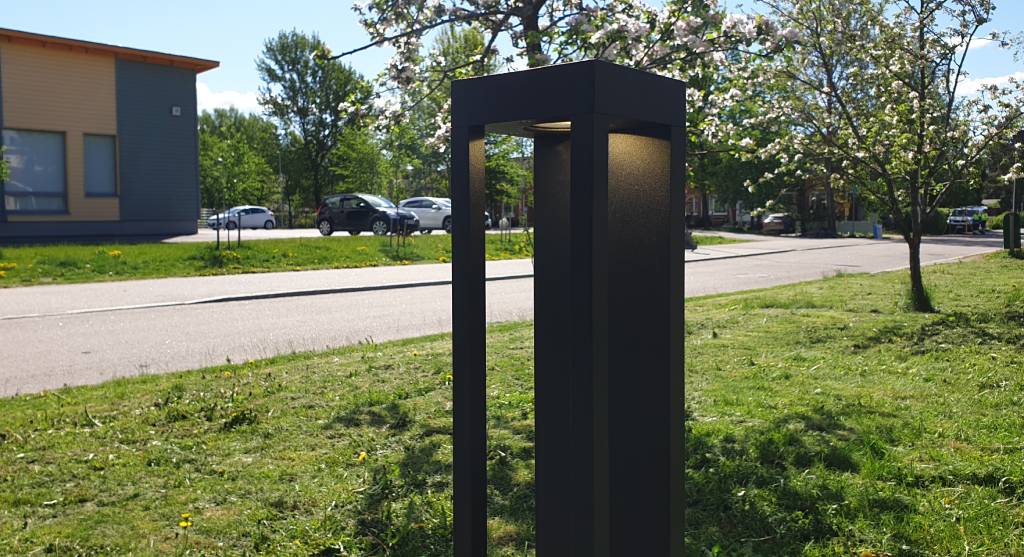 Tyylikäs LED-ulkovalaisin seisoo nurmialueella lähellä jalkakäytävää ja tietä. Kukkivat puut ja etäällä siintävät rakennukset luovat upean taustan kirkkaalle siniselle taivaalle. Täydellinen valaisuratkaisu LedStore.fi -verkkokaupan valikoimasta, joka nostaa pihasi valaistuksen uudelle tasolle.