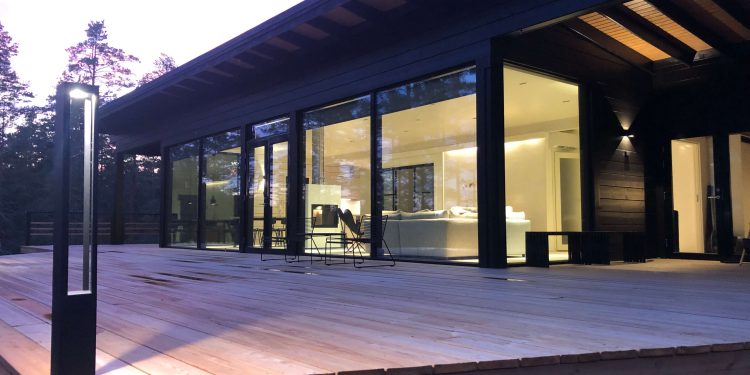 Moderni talo, jossa på suuret lasiseinät ja laaja puinen terassi. LedStore.fi tarjoaa sisätilan LED-valaistuksen, joka valaisee tyylikkäät huonekalut. Iltaisin taivaan ja ympäröivien puiden heijastukset luovat lasipintoihin kauniita yksityiskohtia, jotka viimeistelevät talon modernin ilmeen.