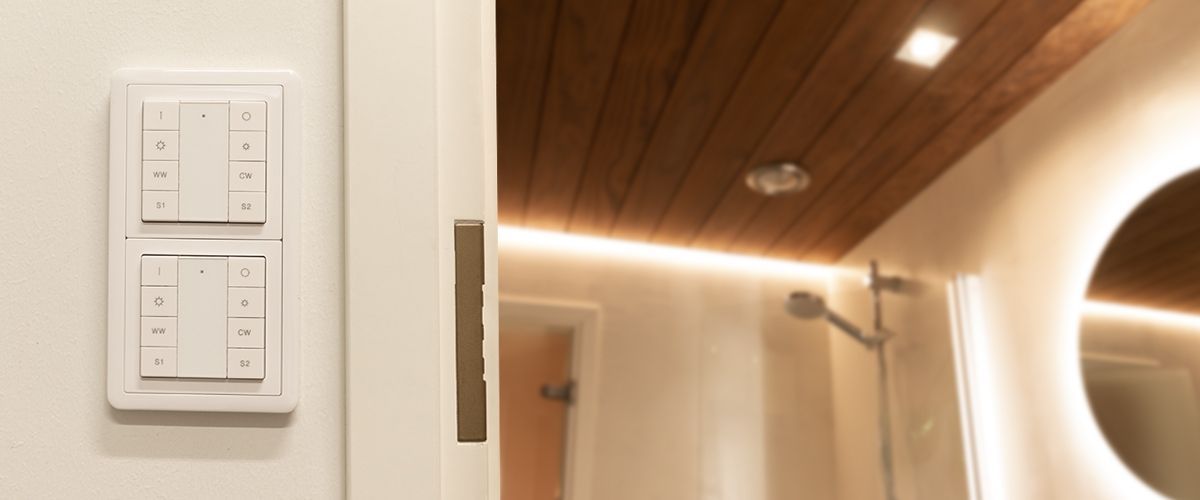 LedStore.fi-verkkokaupassa esitellään moderni kylpyhuone, jossa on puinen katto ja upotettu LED-valaistus, joka korostaa pyöreää peiliä. Ovi on hieman raollaan, paljastaen seinällä olevan digitaalisen ohjauspaneelin. Taustalla näkyy suihkualue.